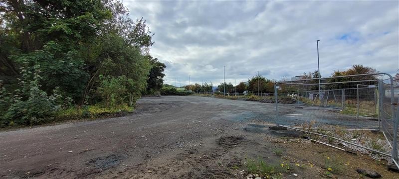 Image showing land which is mostly flat, dirt and gravel track with trees to the left and metal fence barriers on the right